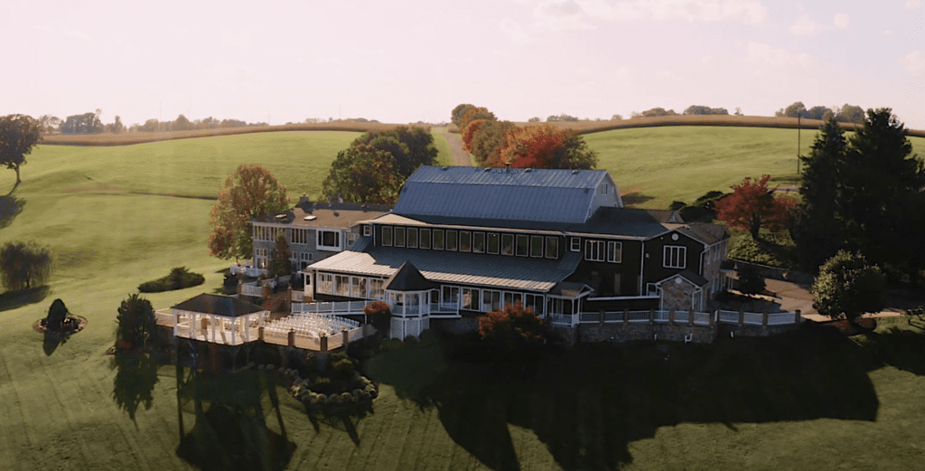 Aerial view of Morningside Inn with multiple sections, nestled in a lush, green landscape. Surrounded by trees with autumn foliage, the house features a deck and expansive windows. Rolling hills stretch into the distance under a clear sky. | Morningside Inn